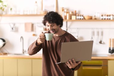 Handsome young man sips coffee and engages with his laptop in a warm kitchen atmosphere.