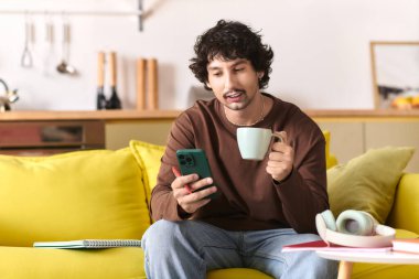 A handsome young man enjoys coffee and checks his phone on a cozy yellow couch.