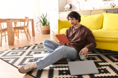 Handsome young man sits comfortably on the rug, reading a book and sipping a drink at home.
