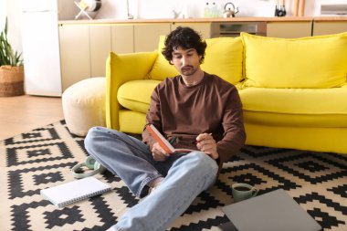 Handsome young man sits on a rug, reading quietly in his chic living room space.