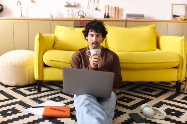 A handsome young man relaxes on the floor with coffee, focused on his laptop in a stylish room.