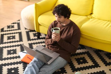 Handsome young man sits on floor with laptop and coffee, surrounded by a bright and comfy space.