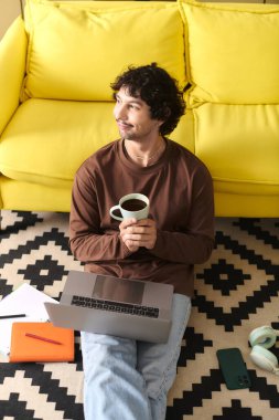 A young, handsome man smiles on the floor with a coffee mug while working on his laptop.
