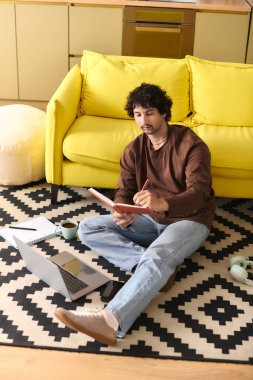 A young man sits on the floor, surrounded by a vibrant yellow couch and warm decor, sketching ideas.