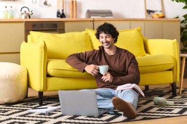 A young man sits comfortably on the floor, enjoying a drink while working on his laptop.