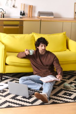A young man sits comfortably on the floor, sipping coffee and taking notes while studying.