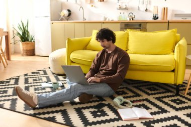 Handsome man sits on the floor, focused on laptop in a warm home setting with lively decor.