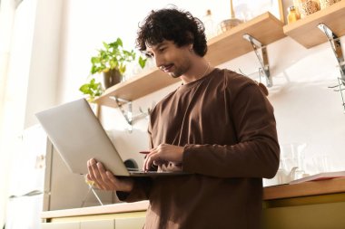 A young man with curly hair smiles while using his laptop in a modern kitchen