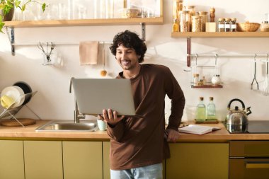 A young man stands in his modern kitchen, smiling as he uses his laptop.