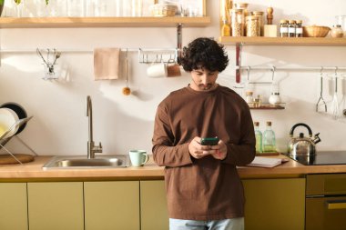 A handsome young man stands in a stylish kitchen, focused on his phone during the day.