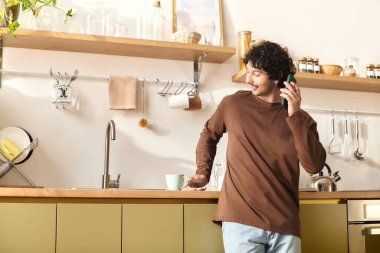 Handsome young man chats on the phone while standing in a modern kitchen.