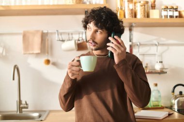 A young, handsome man talks on the phone while sipping coffee in a modern kitchen, looking engaged.