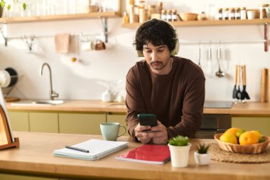 A handsome young man sips coffee in a bright kitchen filled with fruits while on his phone.