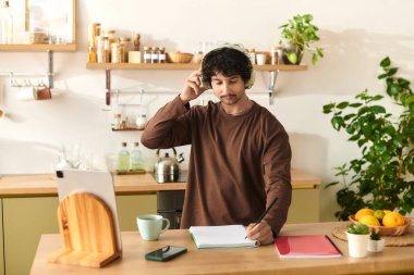 A handsome young man with headphones is focused on note taking at his kitchen desk.