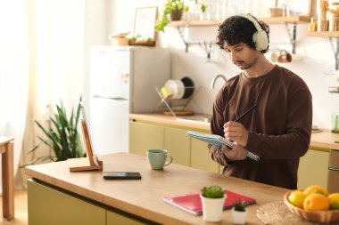 Handsome young man wearing headphones writes notes at a kitchen table filled with fruits and plants.