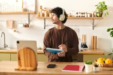 Young man with headphones takes notes in a vibrant kitchen filled with fresh ingredients.
