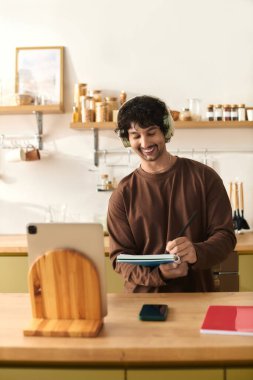 Smiling young man is writing in a notebook while listening to music in a modern kitchen.