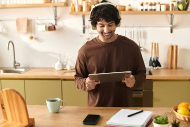 A cheerful young man in a cozy kitchen enjoys using a tablet while sipping coffee.