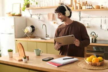 Young man with headphones smiles while using a tablet in his warm kitchen environment.
