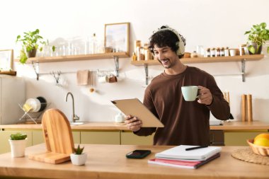 Handsome young man smiles while holding a coffee cup and using a tablet in his home kitchen.
