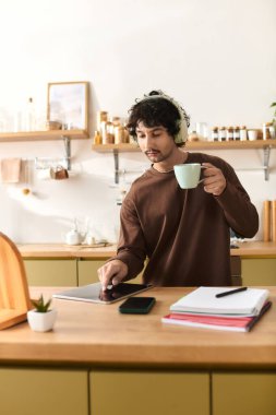 A handsome young man sips coffee at the kitchen counter, focused on his tablet.