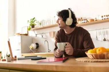 Young man sits in a modern kitchen, sipping coffee and listening to music with headphones on.