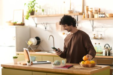 A young man smiles in a cozy kitchen, checking his phone among fresh fruit.