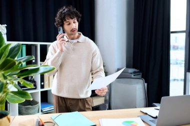 Young man in a stylish sweater talks on the phone while reviewing documents at work.