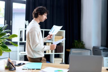 Handsome young man reviews charts and documents at his desk, focused on his work tasks.