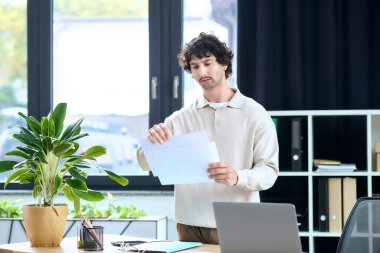 Handsome young man stands in a modern office, examining important papers with focus and intention.