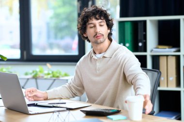 Handsome young man sits at a sleek desk, focused on work while reaching for a coffee cup.