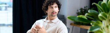 Handsome young man relaxed in a stylish cafe while sipping a hot drink, surrounded by greenery.