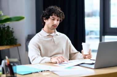 A handsome young man studies charts and reports at his desk while enjoying a cup of coffee.