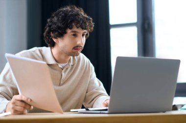 Handsome young man sits at a wooden desk, working intently on a laptop with documents in hand.