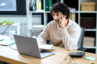 A young man with curly hair sits thoughtfully at his desk, working diligently on his laptop.