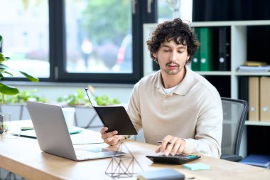 A young man with curly hair is focused on his work at a desk, using a laptop and notebook.