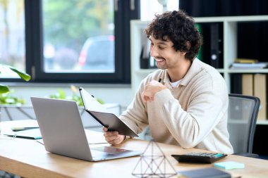 A young man smiles while checking notes on his tablet during a productive work session at his desk.