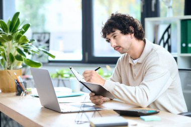 Focused young man writes in a notebook while working on his laptop at a modern office desk.