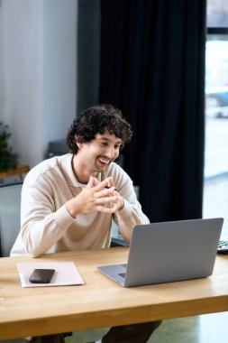 A cheerful young man smiles at his laptop, immersed in a video call from a modern workspace.