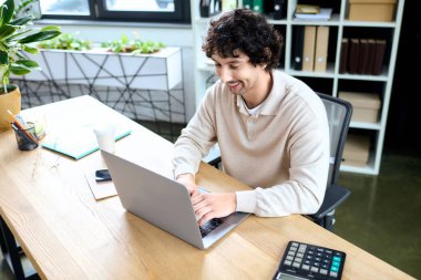 Handsome young man types on a laptop, surrounded by greenery in a stylish workspace.
