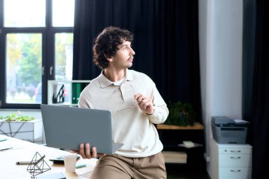 Handsome young man holds a laptop while thinking deeply in a stylish office space.