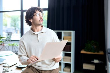 Handsome young man sits in a stylish office, pondering over a document in his hands.