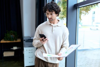 A stylish young man stands in a contemporary office, glancing at his phone while holding papers.