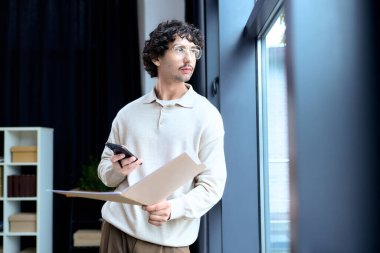 Handsome young man stands near a window, gazing outside while holding a folder and a phone.