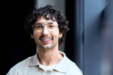 Handsome young man with curly hair and glasses engages with the camera in a bright space.