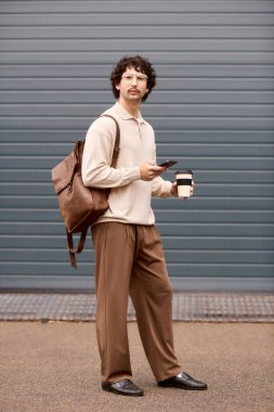 A young man stands confidently with a coffee cup and phone, dressed in stylish casual wear.