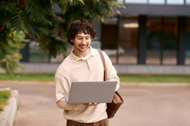 A young man with curly hair enjoys working on his laptop outdoors, surrounded by greenery.