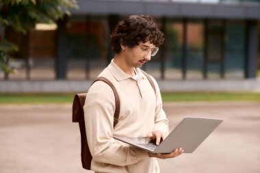 Young man with glasses works on his laptop outside a modern building.