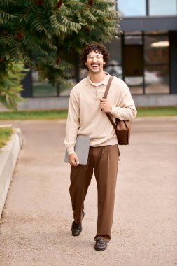 A handsome young man walks confidently on a sunny day, carrying a laptop and bag while smiling.