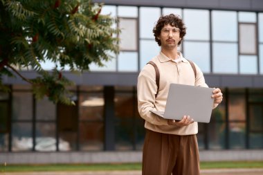 A handsome young man in a cozy sweater holds a laptop in a bright outdoor setting.